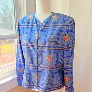 Puede incluir: Una chaqueta azul con un estampado floral con rosas rosas y un dise&ntilde;o repetido de marcos ornamentados. La chaqueta tiene cuello en V y cierre de bot&oacute;n.