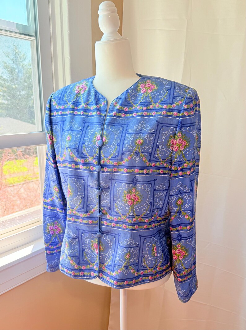Puede incluir: Una chaqueta azul con un estampado floral con rosas rosas y un dise&ntilde;o repetido de marcos ornamentados. La chaqueta tiene cuello en V y cierre de bot&oacute;n.