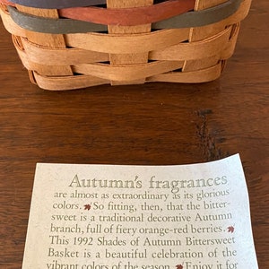 Pode incluir: Cesta tecida com faixas de cores, incluindo marrom, vermelho e verde. A cesta &eacute; feita de material tecido de cor natural. Um cart&atilde;o com texto sobre o design da cesta e a empresa Longaberger est&aacute; em primeiro plano.