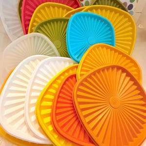 May include: A collection of colorful, square-shaped plastic lids in various shades, including white, red, yellow, green, blue, and orange. The lids have a ridged, fan-like design radiating from the center. The lids are stacked and arranged, showcasing their vibrant colors.