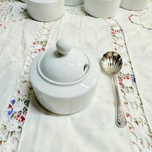 May include: White ceramic sugar bowl with a lid and a silver sugar spoon on a white lace tablecloth.