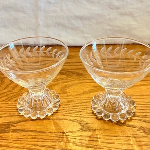 May include: Two clear glass dessert dishes with a delicate etched leaf design around the rim. The dishes have a pedestal base with a beaded edge.