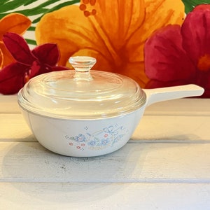 May include: White casserole dish with a floral design and a clear glass lid. The dish has a handle on one side.