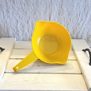 May include: A bright yellow plastic colander with a handle. The colander has a round bowl shape with small holes for draining. The handle has a hole at the end for hanging. The colander is sitting on a white wooden tray.