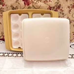 May include: A vintage yellow and white plastic egg container with a lid. The container has a rectangular shape with a compartment for eggs and a separate compartment. The lid is a solid white color with a circular indentation.