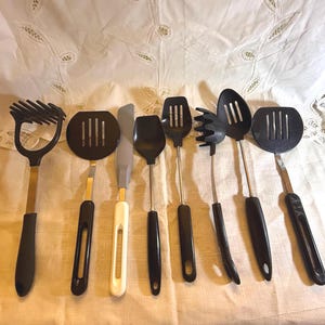 May include: A collection of black kitchen utensils with stainless steel handles. The set includes a potato masher, slotted spatula, solid spatula, spoon, slotted spoon, pasta fork, and a knife-like spatula. The utensils are arranged on a white tablecloth.