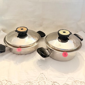 May include: Two stainless steel pots with lids. Each pot has a black handle and a black knob on the lid. The lids are domed and have a circular design on top. The pots are sitting on a white surface.