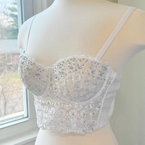 May include: White bustier top with delicate lace and sparkling embellishments. The bodice features a sweetheart neckline and thin, adjustable straps. The design includes intricate beadwork and a scalloped edge, creating a romantic and elegant look.