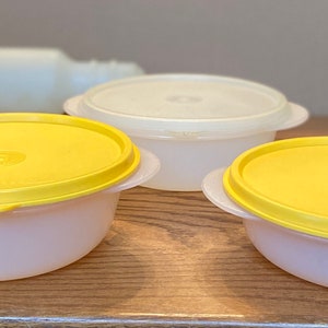 May include: Three round plastic containers with lids. Two containers have yellow lids and one has a white lid. The containers are on a wooden surface.
