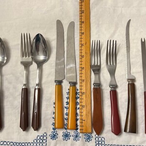 May include: A set of vintage cutlery with brown, red, and yellow plastic handles. The set includes forks, spoons, and knives.