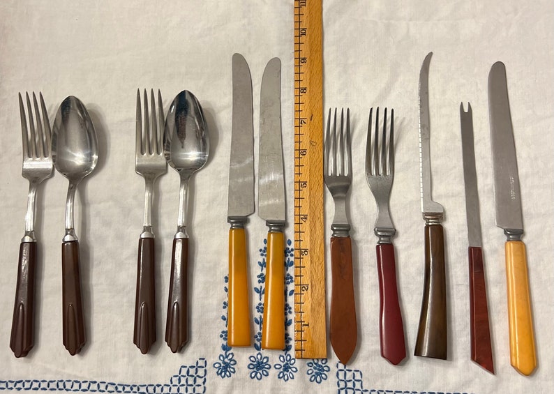 May include: A set of vintage cutlery with brown, red, and yellow plastic handles. The set includes forks, spoons, and knives.