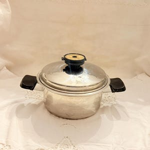 May include: Stainless steel cooking pot with a lid and black handles. The pot has a shiny, reflective surface and a black knob on the lid. The pot is sitting on a white surface.