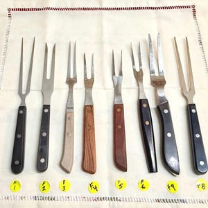 May include: An assortment of vintage carving forks with various handle designs. The forks have stainless steel tines and handles made of wood and black plastic. Each fork is labeled with a yellow sticker.