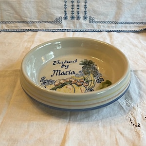 May include: A white ceramic bowl with a blue and pink rim. The bowl has a painted design of fruit and the words "Baked by Maria".