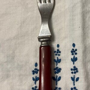 May include: A silver fork with a red plastic handle. The fork has four tines and a simple design.