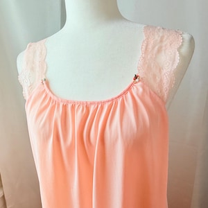 May include: A peach-colored nightgown with delicate lace straps. The straps are adorned with small floral embellishments. The neckline is gathered, and the fabric drapes softly. The nightgown is displayed on a mannequin.
