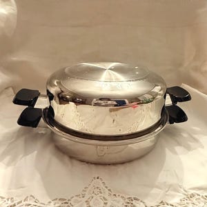 May include: A stainless steel cooking pot with a lid and two black handles. The pot has a shiny, reflective surface and a circular design. The pot is sitting on a white surface.