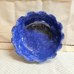 May include: A handmade, blue ceramic bowl with a frilled edge. The bowl has a speckled pattern and a glossy finish. The bowl is likely used for serving food or as a decorative piece.