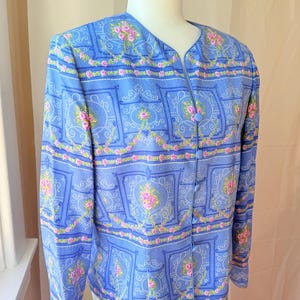 Puede incluir: Una chaqueta azul claro abotonada con un estampado floral con rosas rosas y fondo blanco. La chaqueta tiene cuello en V y mangas largas.