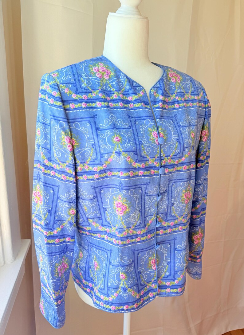 Puede incluir: Una chaqueta azul claro abotonada con un estampado floral con rosas rosas y fondo blanco. La chaqueta tiene cuello en V y mangas largas.