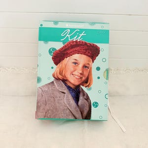 May include: A teal box featuring a portrait of a girl wearing a red beret and gray coat. The box has the words "American Girl" and "Kit 1934" printed on it. The box is decorated with teal circles and swirls.