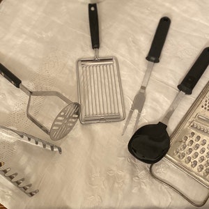May include: A set of five kitchen utensils, including a potato masher, a pair of tongs, a spatula, a fork, and a grater. The utensils are all made of stainless steel and have black plastic handles.