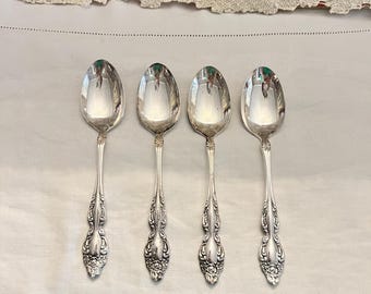 May include: Four silver spoons with ornate handles. The spoons are arranged in a row on a white tablecloth.