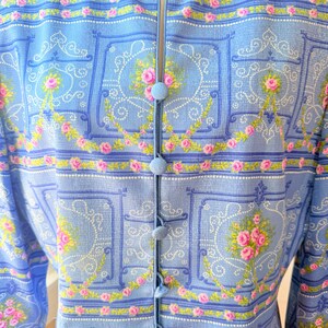 Puede incluir: Una blusa azul claro con botones y estampado floral con rosas rosas y un patr&oacute;n repetido de cuadrados y espirales azules. La blusa tiene cuello en V y mangas largas.