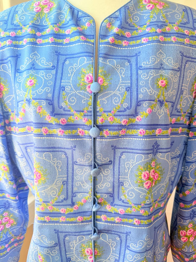 Puede incluir: Una blusa azul claro con botones y estampado floral con rosas rosas y un patr&oacute;n repetido de cuadrados y espirales azules. La blusa tiene cuello en V y mangas largas.