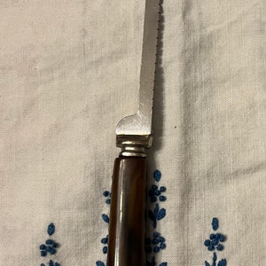 May include: A silver serrated knife with a brown wooden handle. The knife is lying on a white fabric surface with blue embroidered flowers.