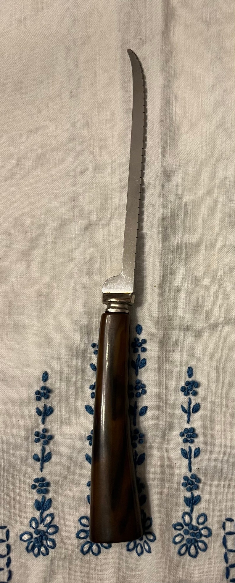 May include: A silver serrated knife with a brown wooden handle. The knife is lying on a white fabric surface with blue embroidered flowers.
