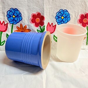 Peut inclure: Un pot à ustensiles en céramique bleu avec des rainures verticales, à côté d'un gobelet en plastique translucide. L'intérieur du pot est beige clair. Le fond est un tissu blanc avec des broderies florales colorées.