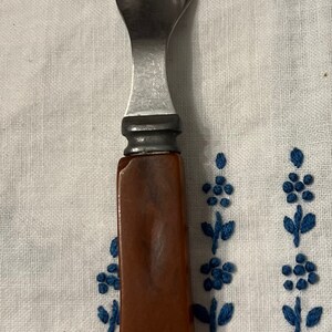 May include: A silver fork with a brown plastic handle. The fork is lying on a white tablecloth with blue embroidered flowers.