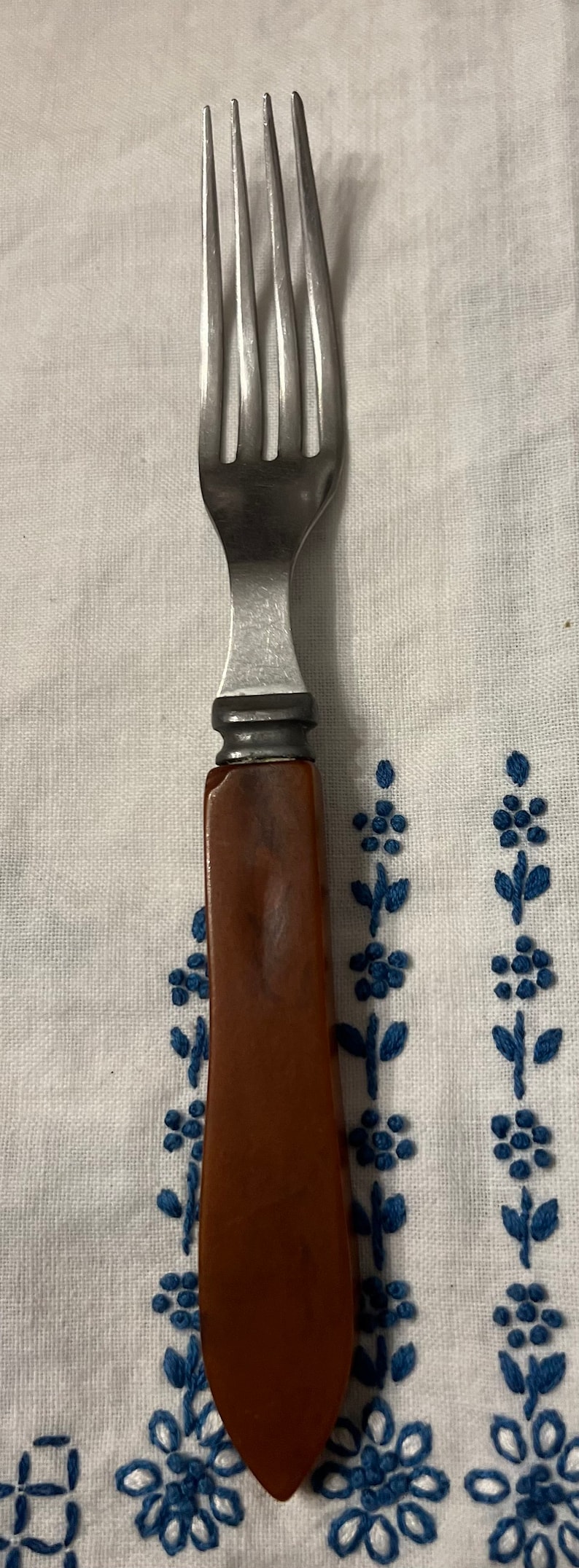 May include: A silver fork with a brown plastic handle. The fork is lying on a white tablecloth with blue embroidered flowers.