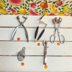 May include: A selection of vintage kitchen utensils, including a nutcracker, cherry pitter, and bottle opener, displayed on a white wooden surface. The tools are made of metal and have different handle designs. A colourful fruit-themed fabric is in the background.