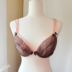 May include: A peach-colored bra featuring a black lace overlay on the cups. The garment includes thin peach straps, a central peach-colored bow, and small black bows at the base of each strap where it connects to the cup, displayed on a mannequin.