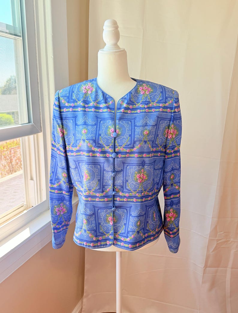 Puede incluir: Una chaqueta azul con un estampado floral con rosas rosas y fondo blanco. La chaqueta tiene cierre de botones y mangas largas.