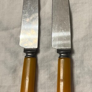May include: Two silver butter knives with yellow plastic handles. The knives have a slightly curved blade and a serrated edge.