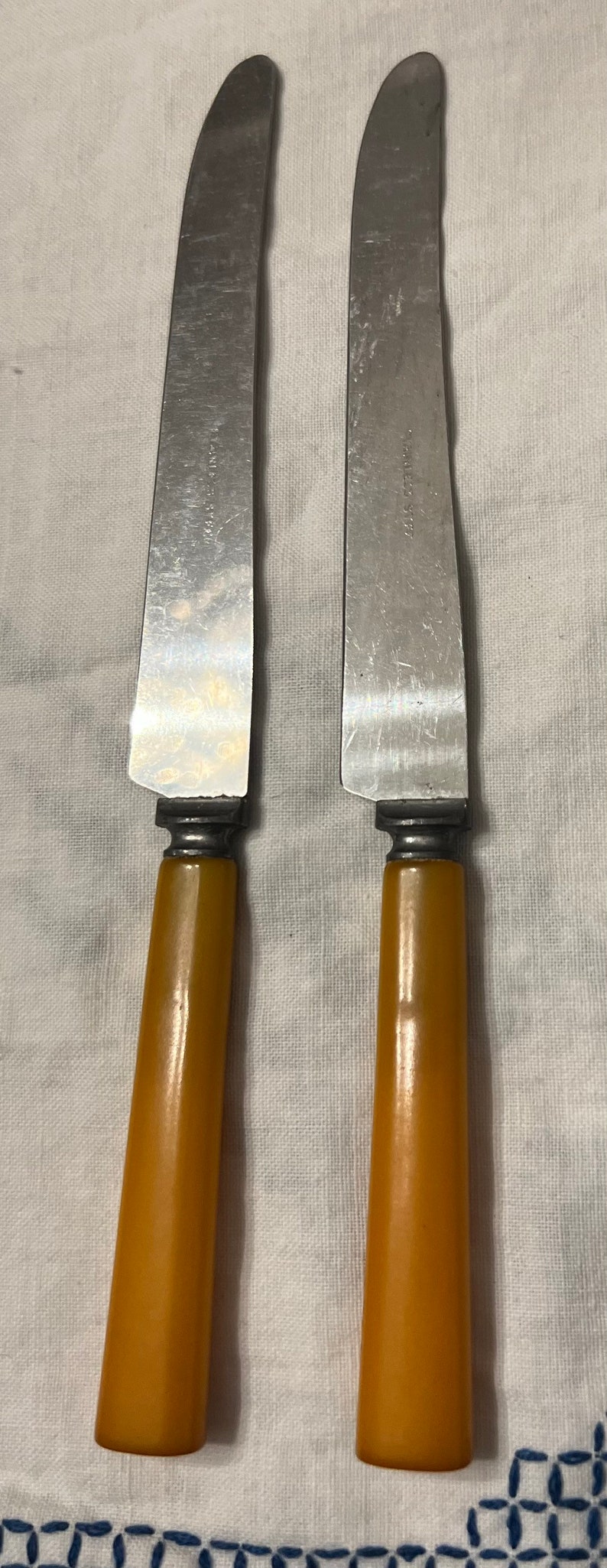 May include: Two silver butter knives with yellow plastic handles. The knives have a slightly curved blade and a serrated edge.