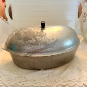 May include: A vintage, oval-shaped aluminum roasting pan with a lid and a black handle. The pan has a slightly textured surface and is sitting on a white lace tablecloth. The lid has a small, round black handle.