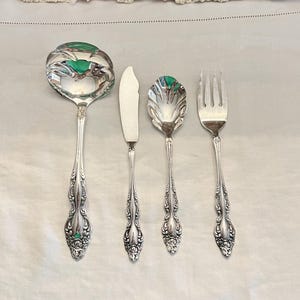 May include: A set of four silver-plated serving utensils, including a ladle, butter knife, sugar spoon, and a fork. The utensils have ornate handles with a floral design.