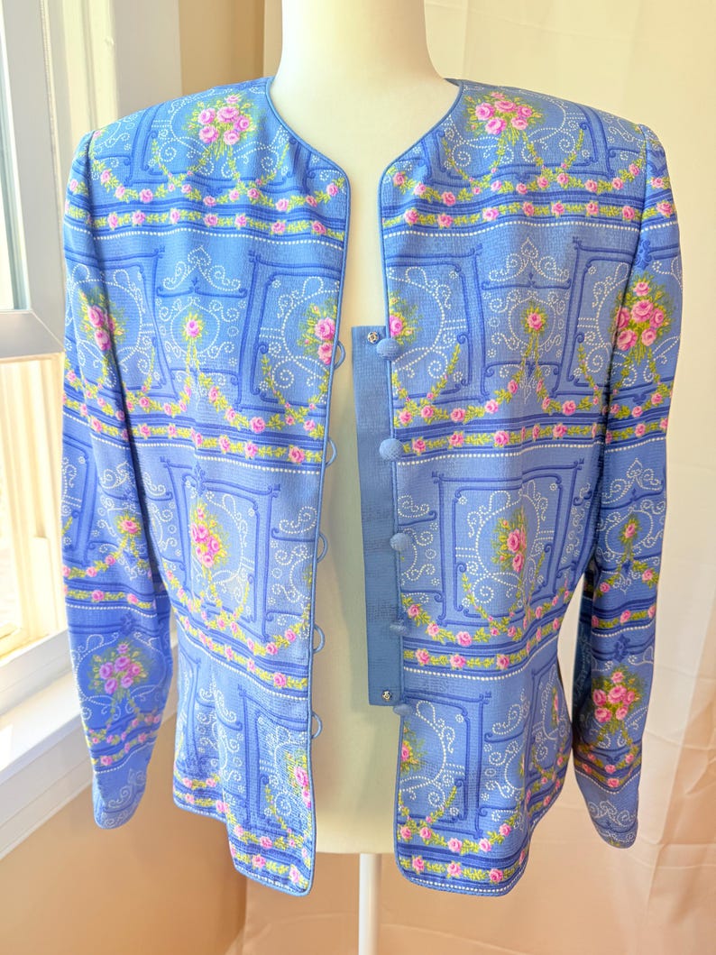 Puede incluir: Una chaqueta azul claro con un estampado floral con rosas rosas y fondo blanco. La chaqueta tiene cierre de botones y mangas largas.