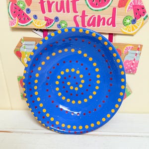 May include: A vibrant blue ceramic plate with a dotted pattern in yellow and red. The plate is round and sits below a sign that reads "fruit stand" with fruit illustrations. The plate has a speckled texture.