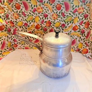 May include: A silver-colored aluminum double boiler pot with a matching lid and two long handles. The pot features a tiered cylindrical body design with a smooth metallic finish and a small black knob centered on the top of the lid.