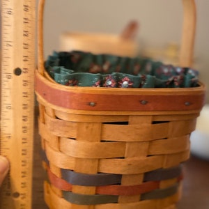 Pode incluir: Uma cesta tecida com uma al&ccedil;a de madeira e forro de tecido decorativo. A cesta tem aproximadamente 20 cm de altura e apresenta faixas vermelhas, azuis e verdes tecidas nas laterais. A borda &eacute; adornada com uma faixa vermelha.