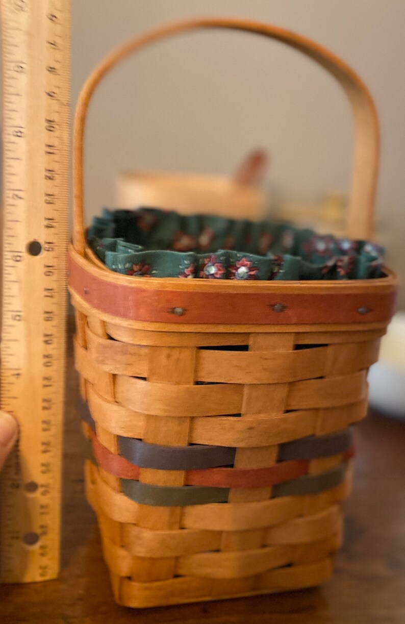 Pode incluir: Uma cesta tecida com uma al&ccedil;a de madeira e forro de tecido decorativo. A cesta tem aproximadamente 20 cm de altura e apresenta faixas vermelhas, azuis e verdes tecidas nas laterais. A borda &eacute; adornada com uma faixa vermelha.