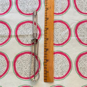 May include: A silver metal fruit peeler with a handle and a blade. The peeler is on a white fabric background with a pattern of pink and green dragon fruit.