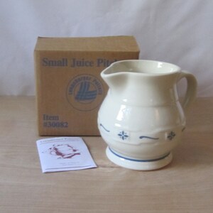 May include: Cream-colored ceramic juice pitcher with a handle and a spout, decorated with blue floral accents and a blue base. A cardboard box labeled "Small Juice Pitcher" and a small card are in the background.