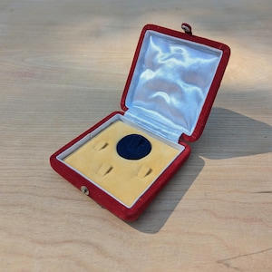 May include: Open red velvet jewelry box with a yellow interior and a dark blue ring holder. The inside lid is lined with white satin. The box is open, revealing the interior, and is set on a wooden surface.