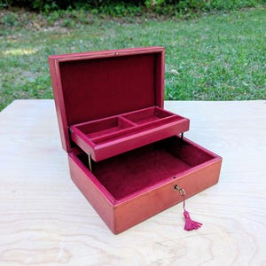 May include: A rectangular red leather jewelry box with a hinged lid and a removable interior tray. The interior is lined with deep red velvet. A small brass lock is attached to the front, featuring a dangling red tassel key for closure.